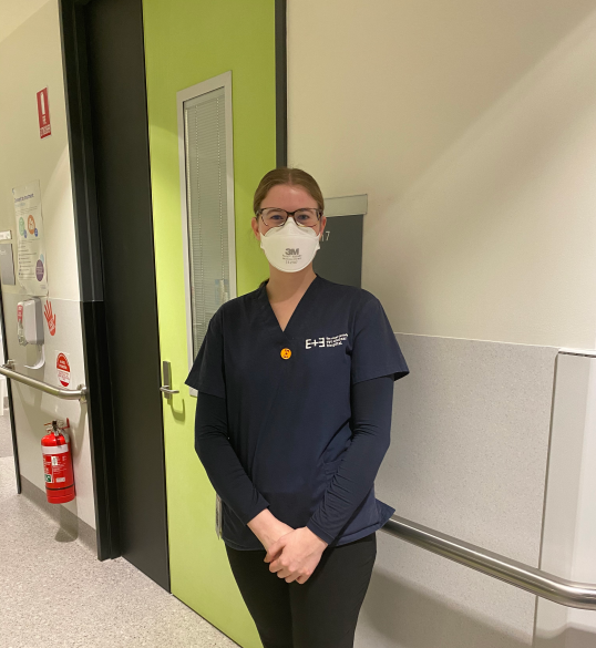 Sarah standing in one of our hospital wards, smiling with an N-95 mask on