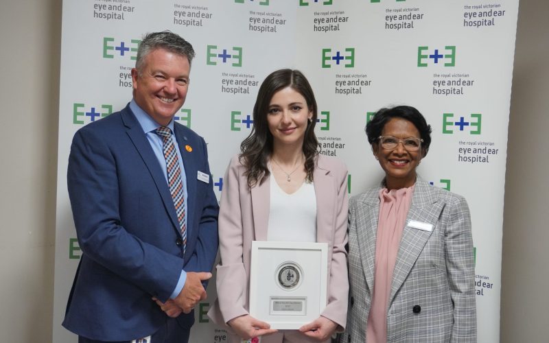 An image of CEO Brendon Gardner and Sherene, with Orthoptist Pelin situated between them. Pelin is wearing a pink suit standing in front of signs with the Eye and Ear logo. She is holding her award that was presented to her at the 2021 Annual General Meeting.