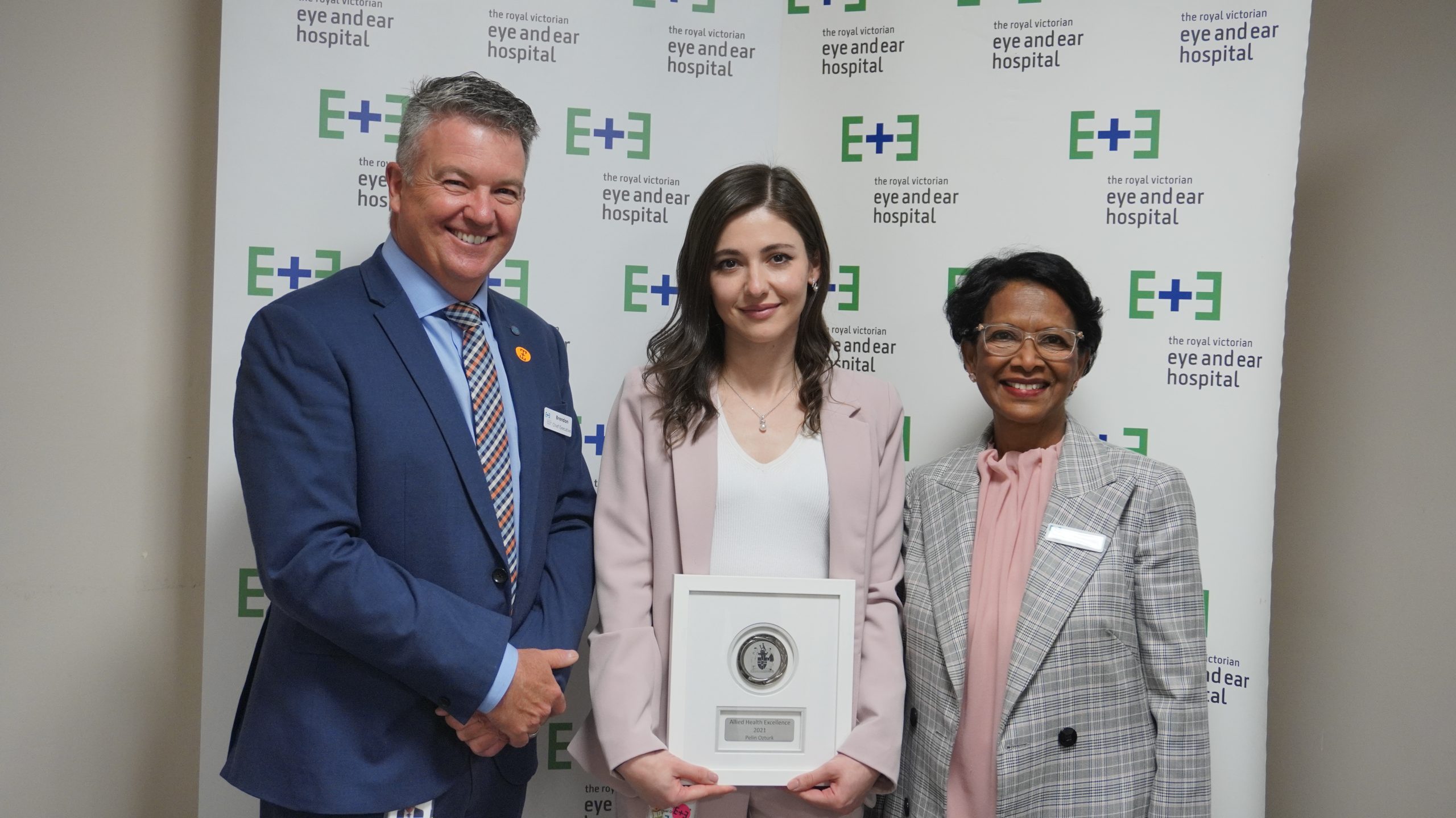 An image of CEO Brendon Gardner and Sherene, with Orthoptist Pelin situated between them. Pelin is wearing a pink suit standing in front of signs with the Eye and Ear logo. She is holding her award that was presented to her at the 2021 Annual General Meeting.