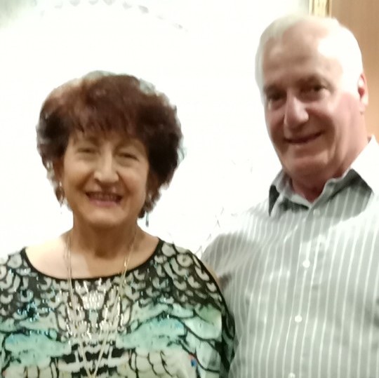 Wife Beatrice (pictured to the left) and Husband John standing and smiling together in front of a white wall.
