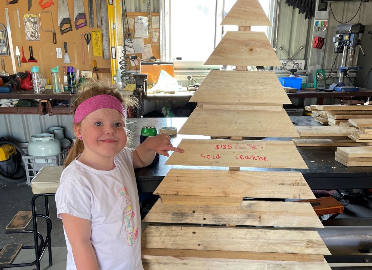 Smiling Katie standing next to one of the wooden christmas trees she made with her grandfather