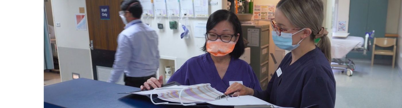 Two nurses wearing scrubs and face masks reviewing a patient chart.