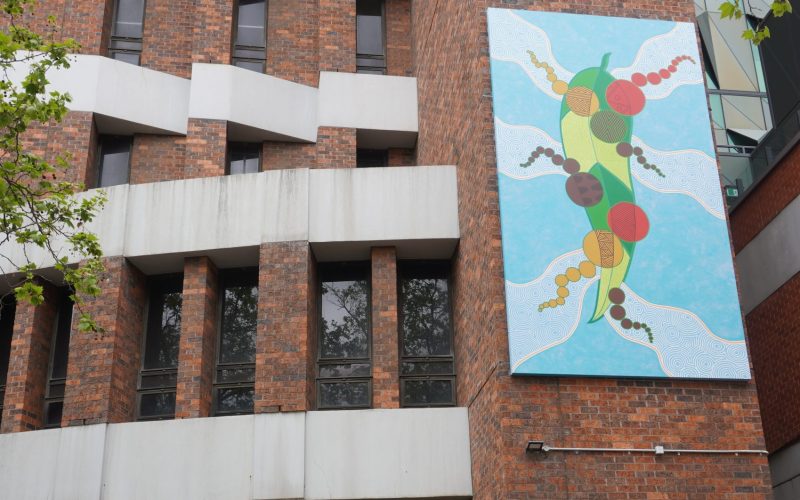 Red and white brick building with colourful painting by Wurundjeri artist Mandy Nicholson turned mural sitting up high.