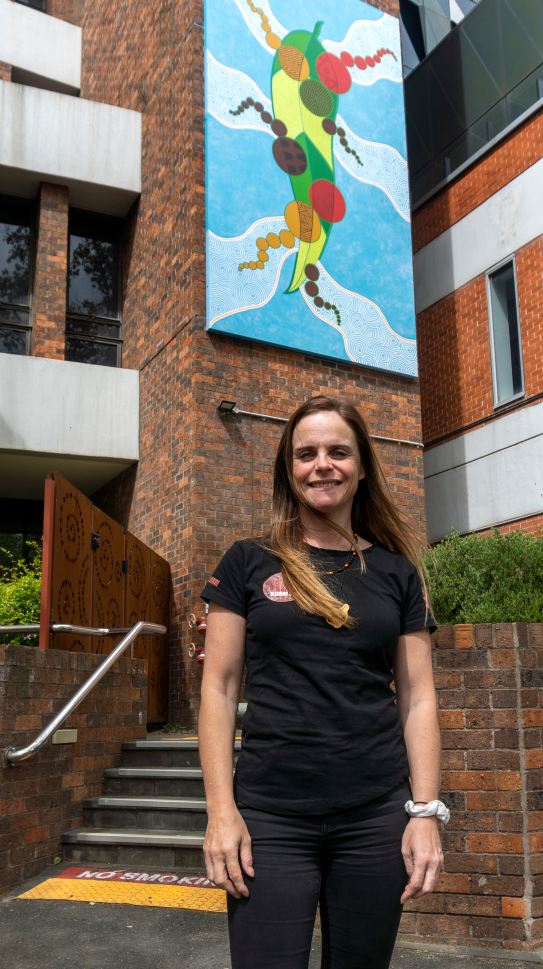Photo of Wurundjeri artist Mandy Nicholson standing in front of her artwork turned mural which is located up high on red brick