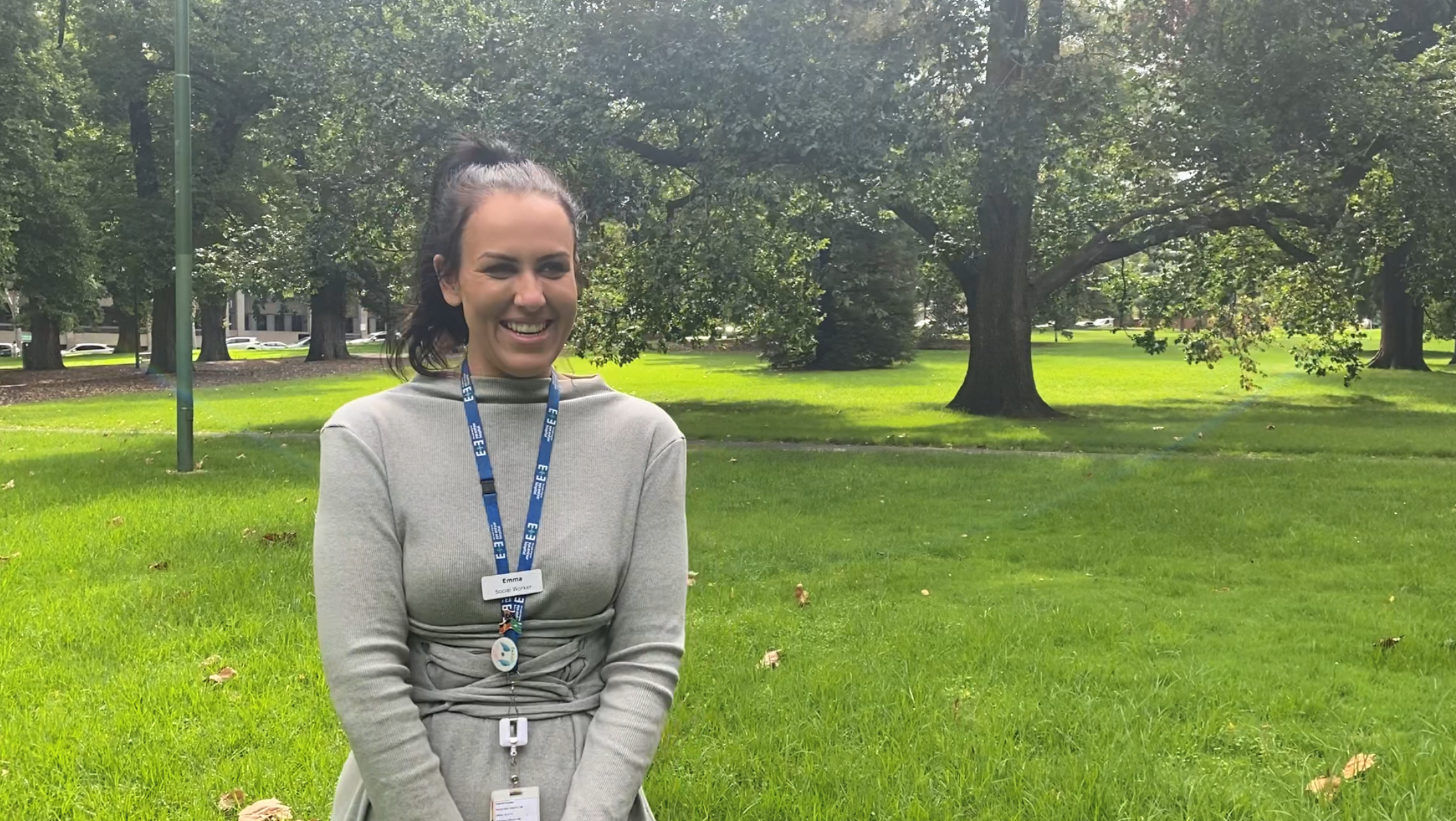 A woman with black hair smiling, behind her is a park with green grass and trees