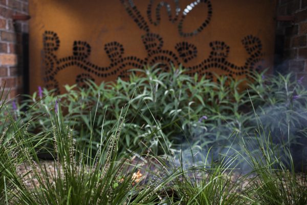 Image of a smoking ceremony bowl in use obscured by bushes, a rust coloured panel depicting aboriginal art is in the background