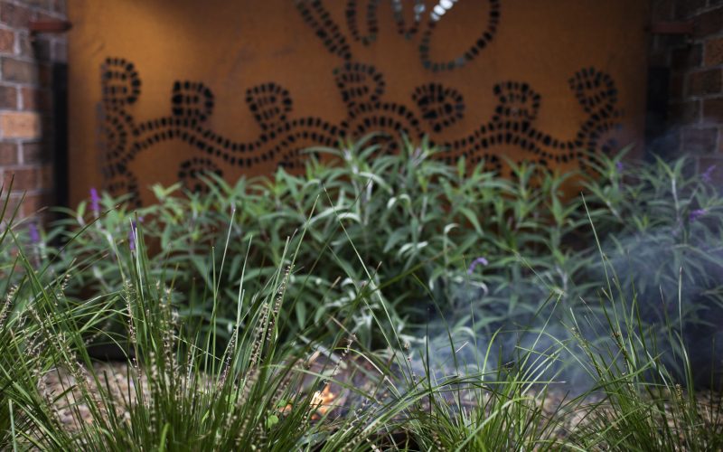 Image of a smoking ceremony bowl in use obscured by bushes, a rust coloured panel depicting aboriginal art is in the background