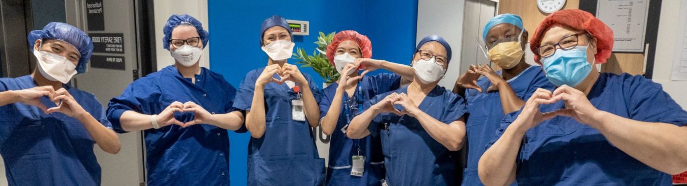 6 nurses wearing blue scrubs and masks, smiling as they make love heart shapes with their hands