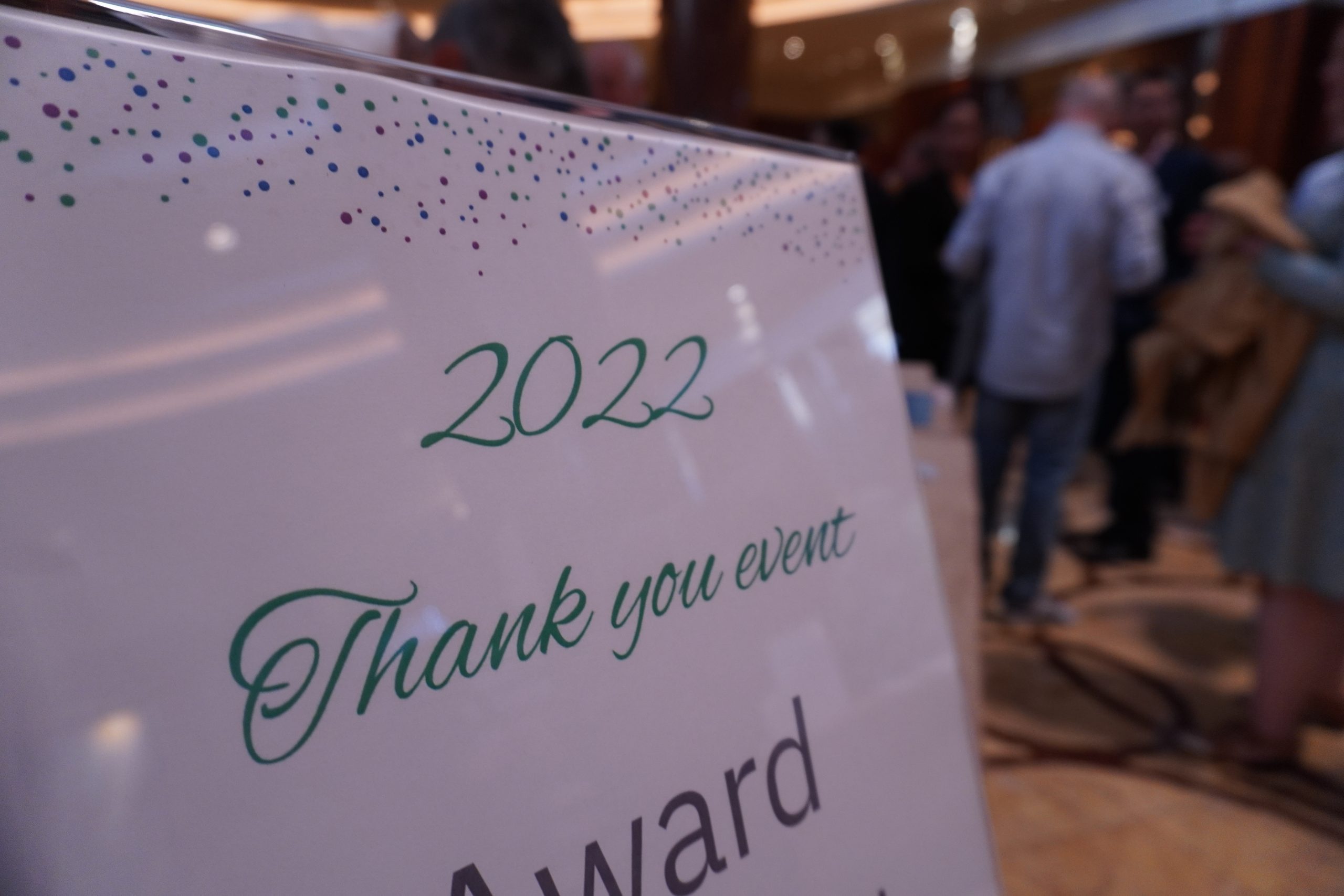 A white lamiated sign saying '2022 Thank you event' in green writing. There is a man with his back to the camera wearing a blue shirt