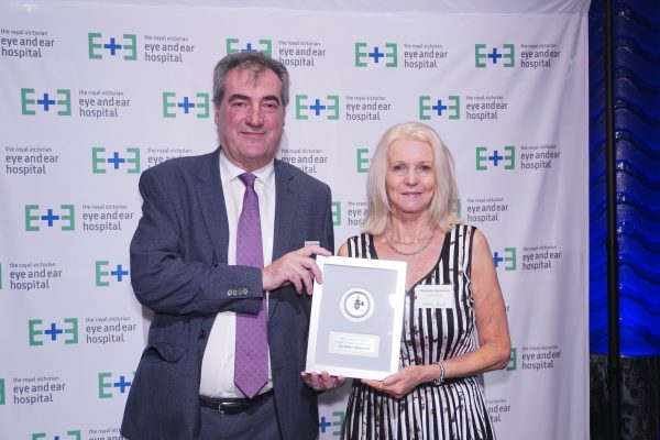 A man with grey hair wearing a greay suit handing over an award to a woman with blonde hair in a black and white dress againsat a white backdrop with the Eye and Ear logo on it