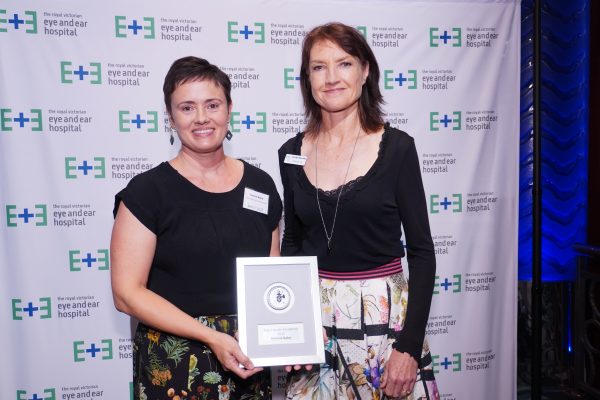 A woman with short black hair and black top holding an award whilst standing next to a woman with brown hair wearing a back top. They're standing against a white backdrop with the Eye and Ear logo on it