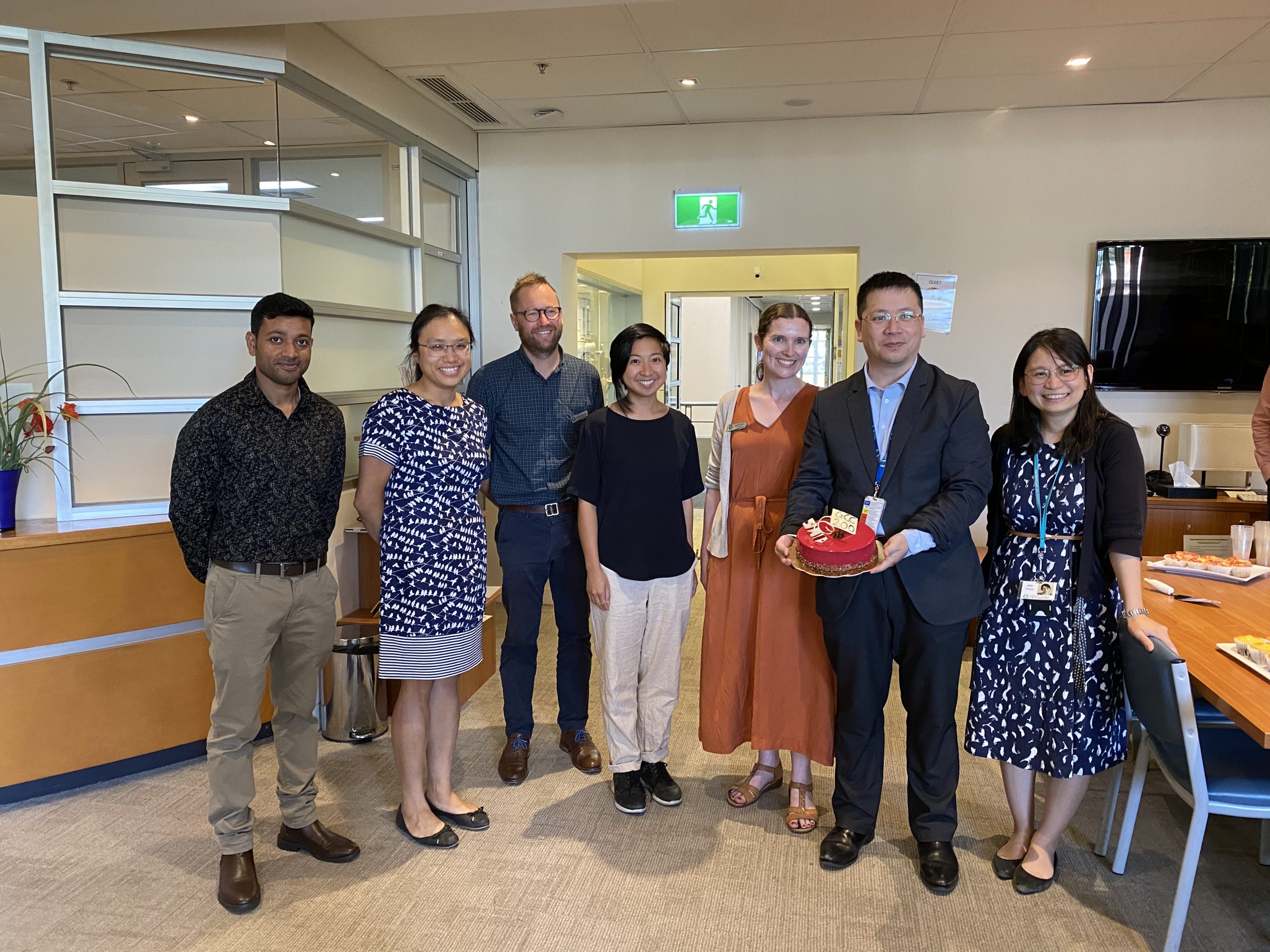 3 men and 4 women standing next to one another indoors, one is holding a red cake that says 'GCC 200'