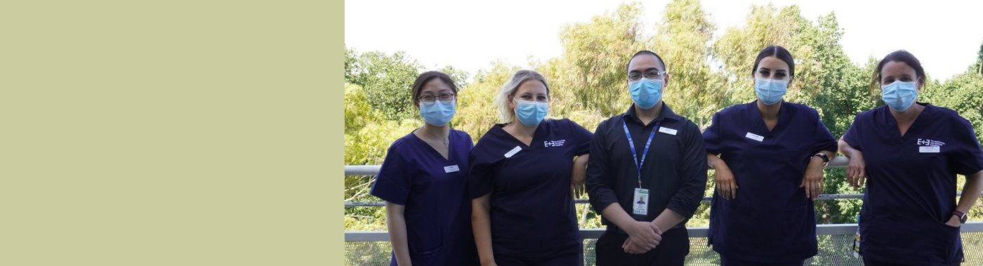 5 nurses standing on a balcony with trees in the background wearing navy scrubs. All are smiling and wearing masks
