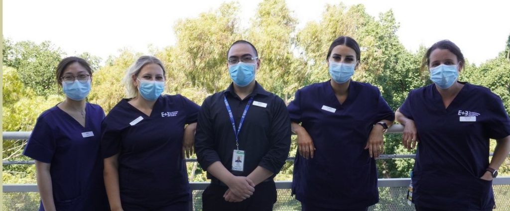 5 nurses standing on a balcony with trees in the background wearing navy scrubs. All are smiling and wearing masks