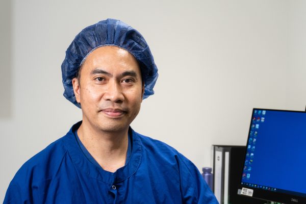 A man wearing a scrub hat and blue scrubs smiling against a white wall with a computer in the background