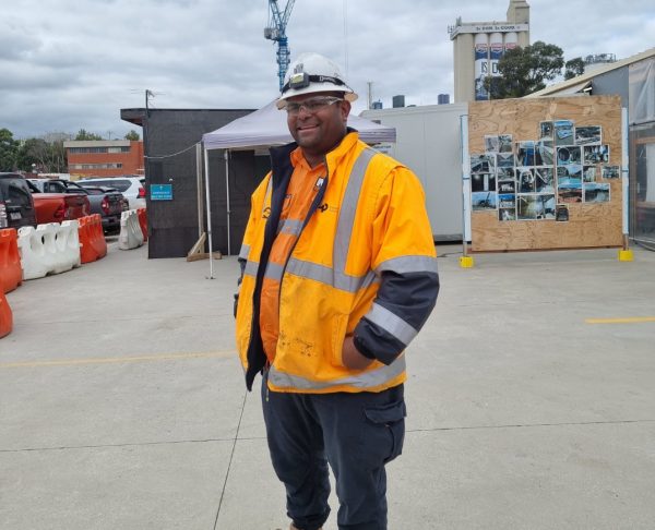 A man standing and wearing an orange hi-vis jacket and builders hat