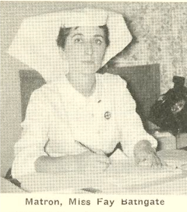 A black and white photo of a woman sitting at a table with a pen in her hand wearing a white nursing top and a a large and broad nursing cap