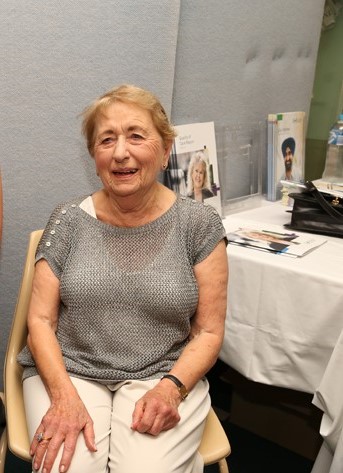 An elderly woman with golden hair in a grey top, sitting down and smiling