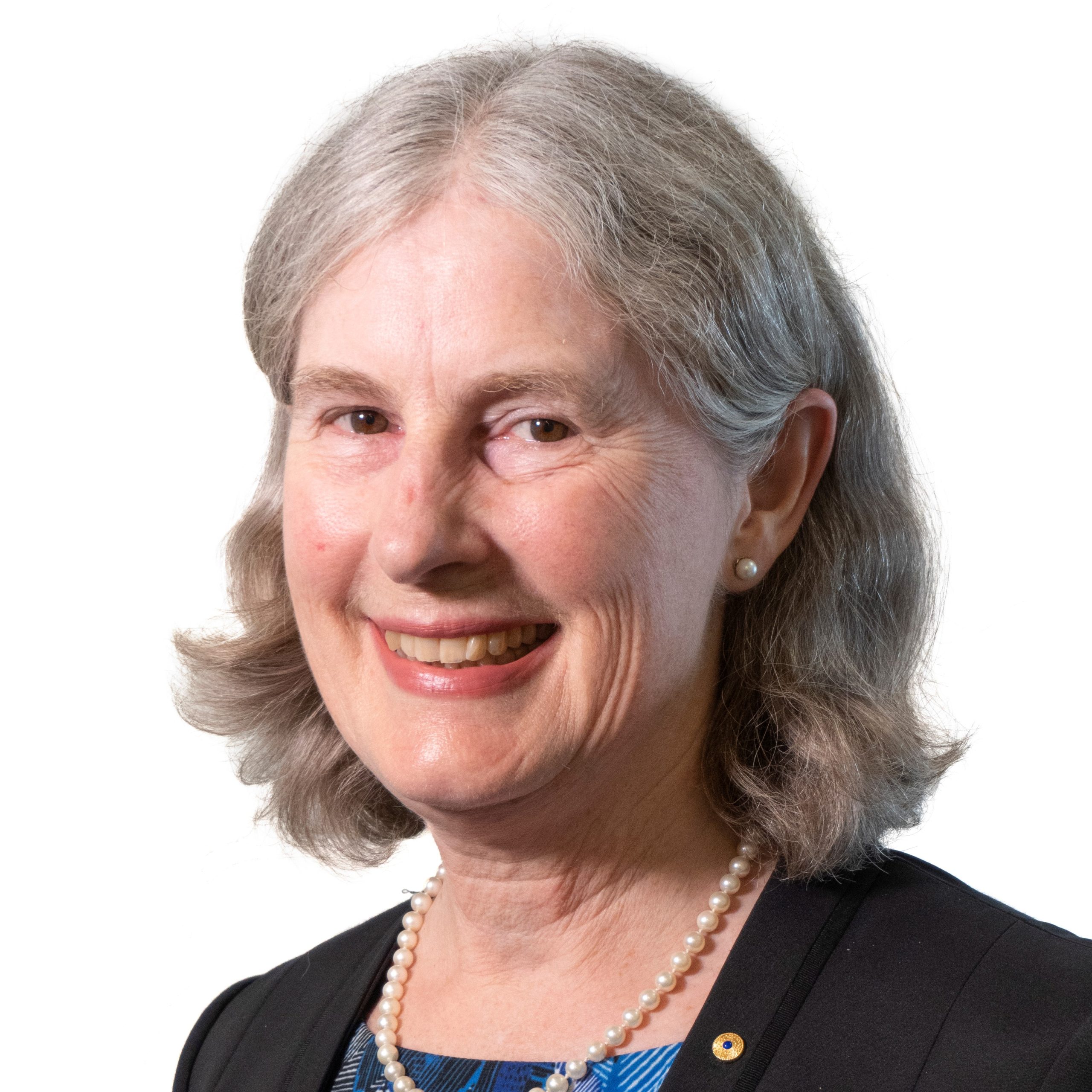 Headshot of a middle-aged female with grey short hair smiling, wearing a dark blue blazer and a light blue delicate shirt and a white pearls necklace.