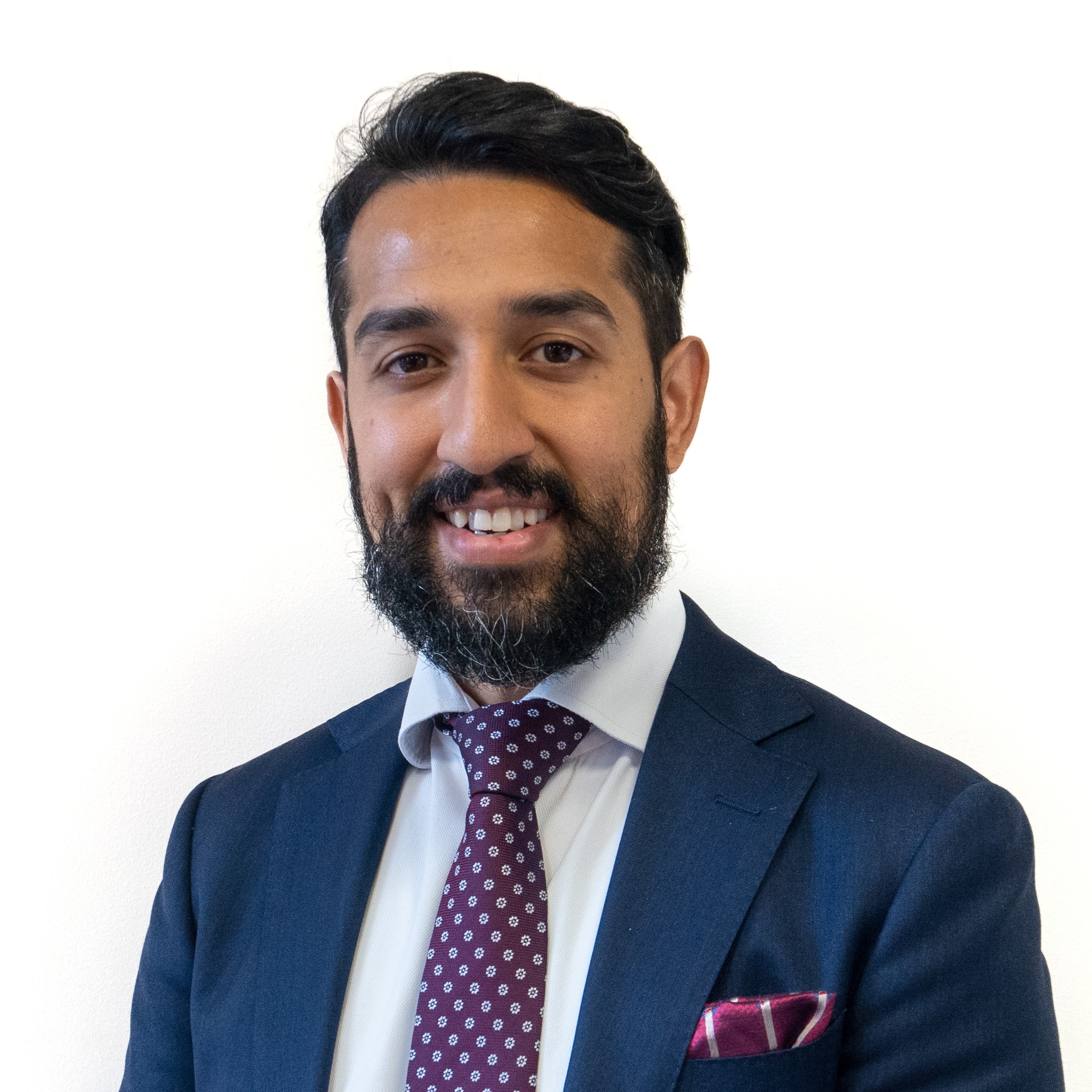 Headshot of a middle-aged male with black short hair and black beard smiling, wearing a dark blue suit, white shirt and purple tie with some white dots on it.