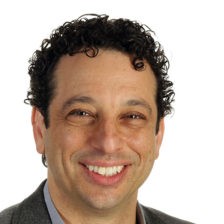 Headshot of a middle-aged male with black short curly hair smiling, wearing a dark grey suit and a grey shirt.