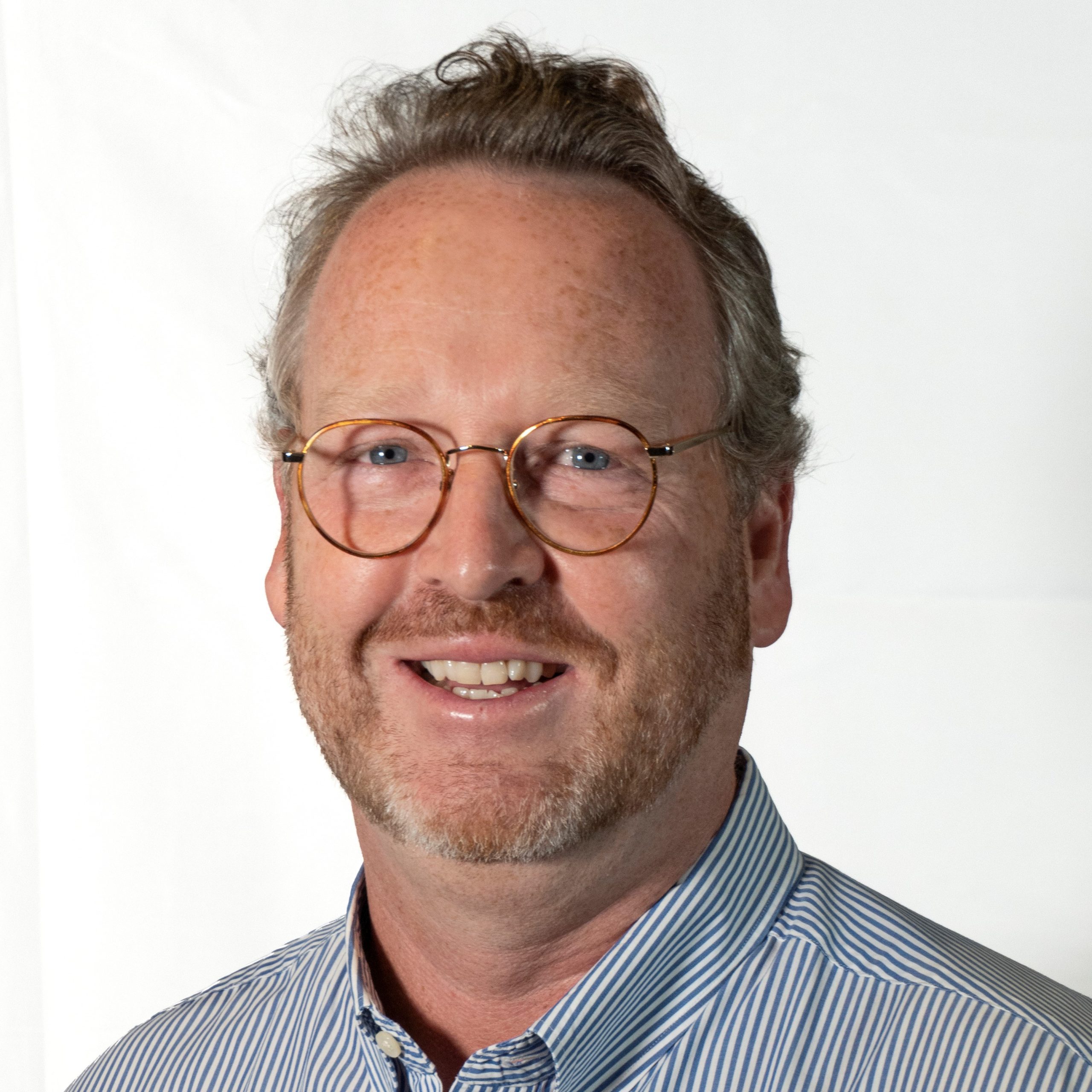 Headshot of middle-aged male with grey short hair smiling, wearing glasses and a light blue shirt with vertical white strips.