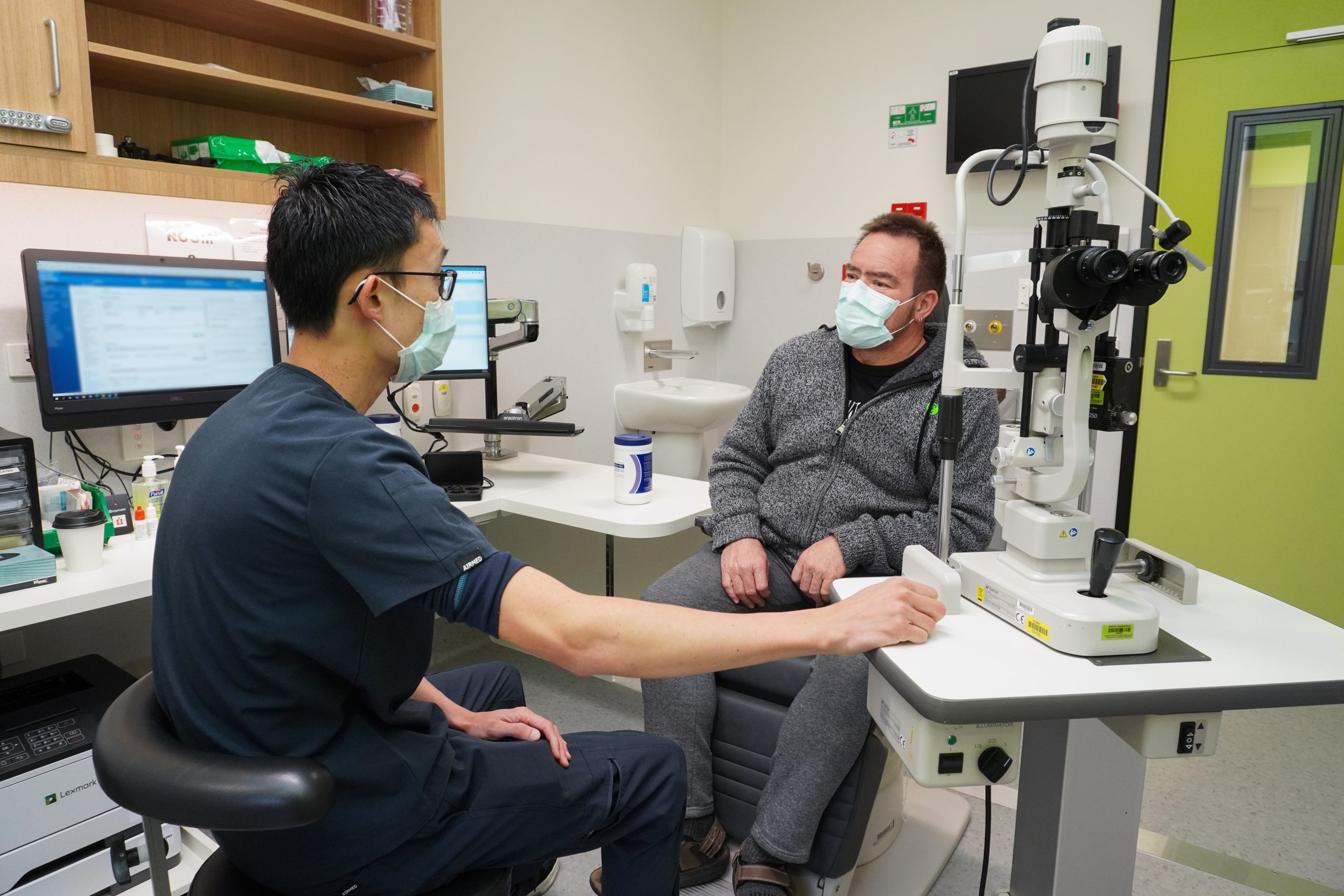 Patient Peter sitting in a chair next to a slit lamp wearing a mask, listening to a doctor