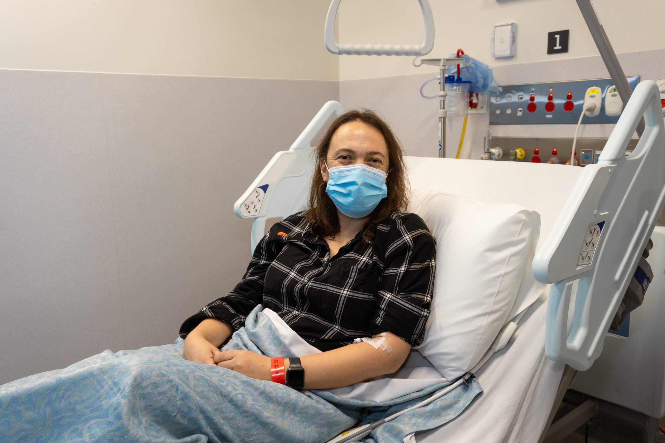 A woman lying in a hospital bed in the new short stay unit, wearing a mask and smiling