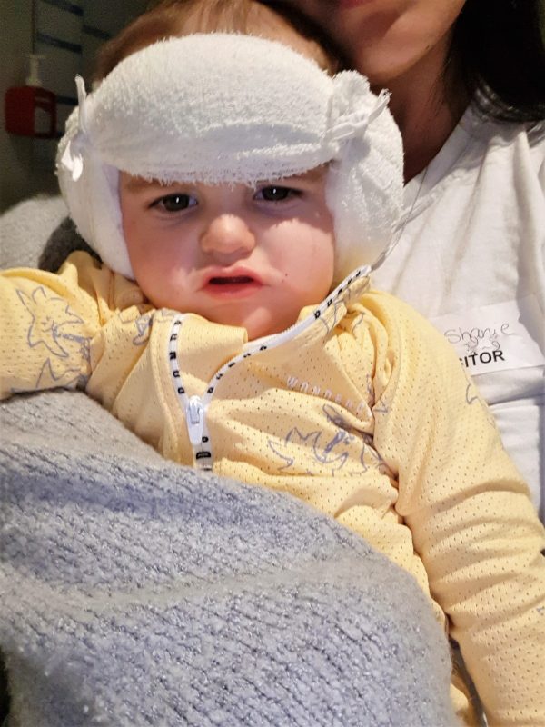 Image of baby know after surgery, his head bandaged and behind held in his mothers arms