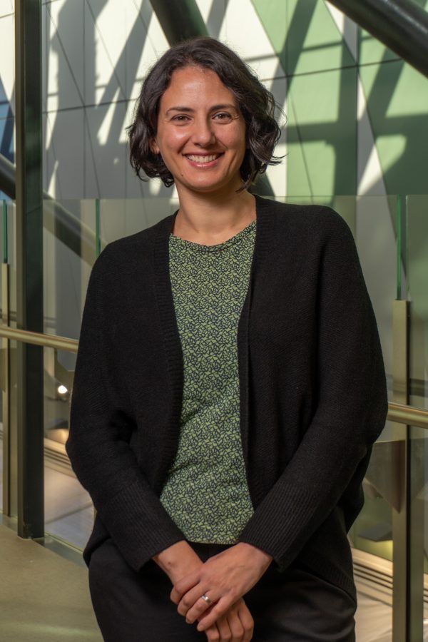 A young woman with short black hair is smiling while wearing a green blouse with black patterns, black blazer and black pants. 