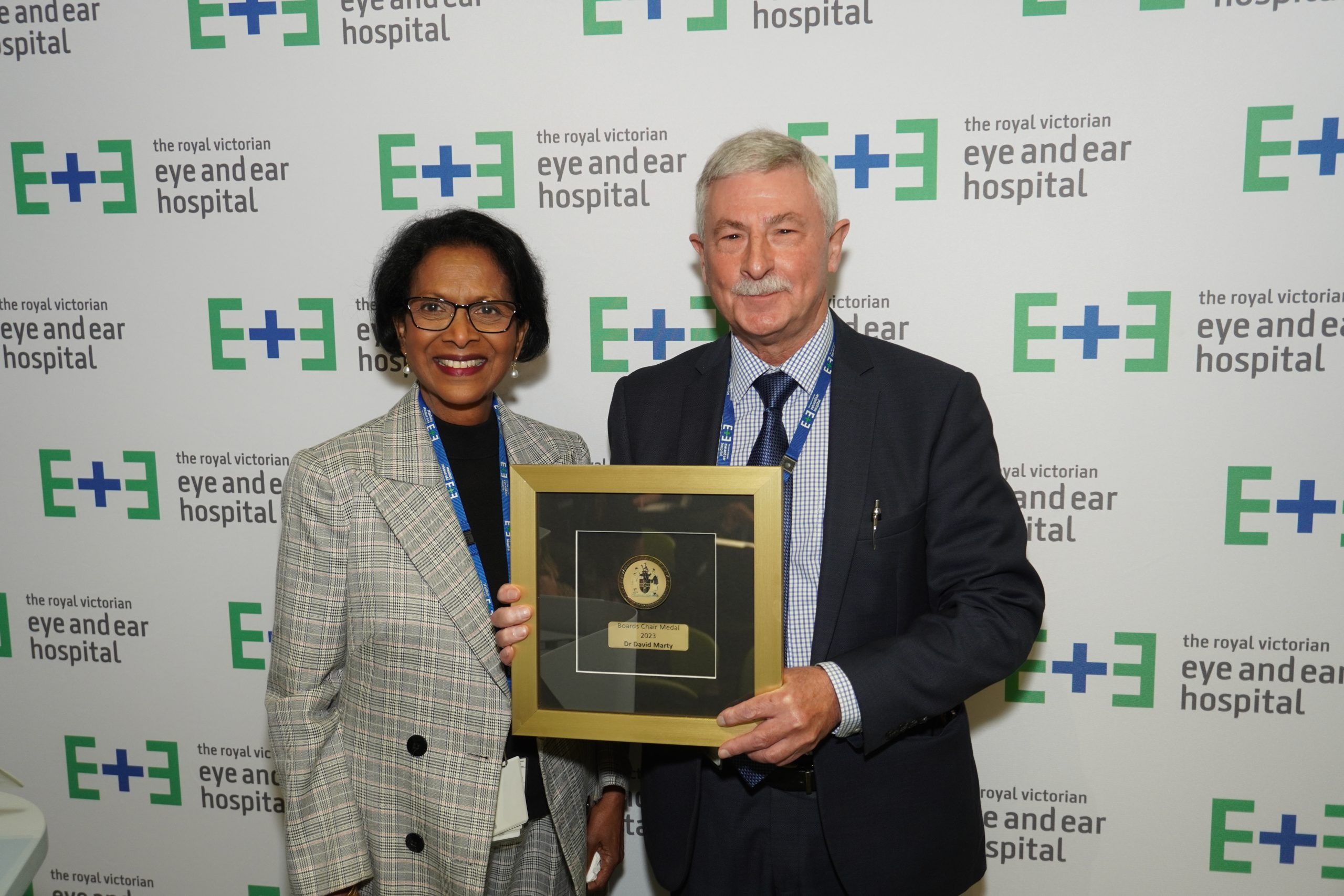 Board Chair Dr Sherene Devanesen standing next to Board Chair Medal Recipient Dr David Marty who is holding the Gold and Black framed award. They stand against an Eye and Ear logo Backdrop.