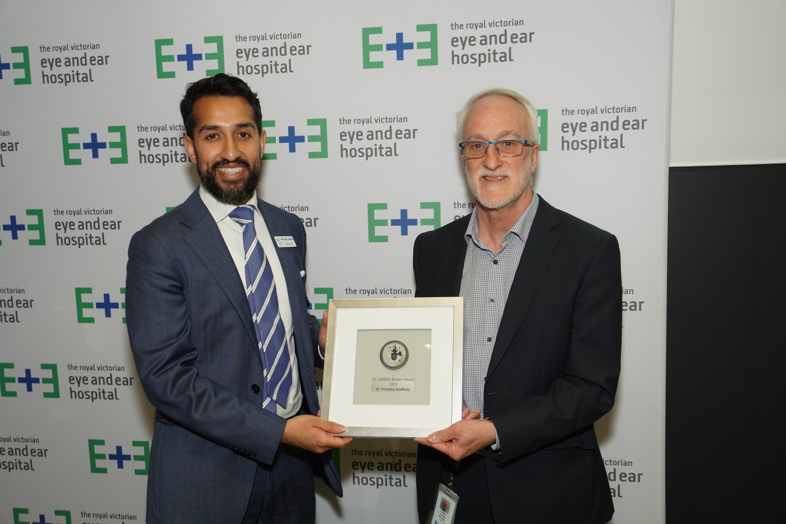 Executive Director Dr Birinder Giddy presenting the Dr J Aubrey Bowen Medal to Dr Tim Godfrey. They stand together smiling against an Eye and Ear logo background