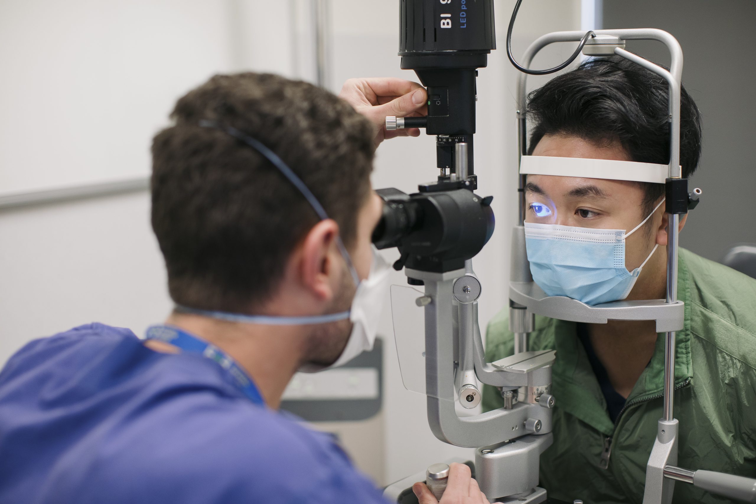 patient having eye examined through slit lamp by a doctor. Both are wearing masks.