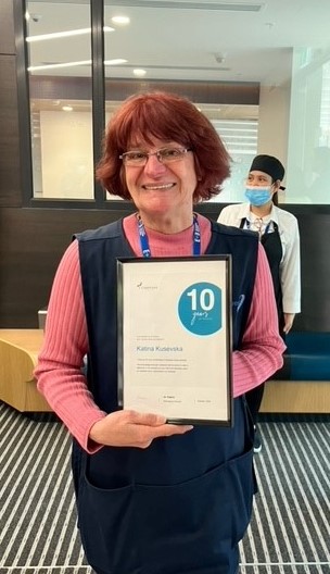 Katina smiling at the camera and holding a 10 year certificate. She wears a pink long sleeve knitted top and a navy cleaning bib