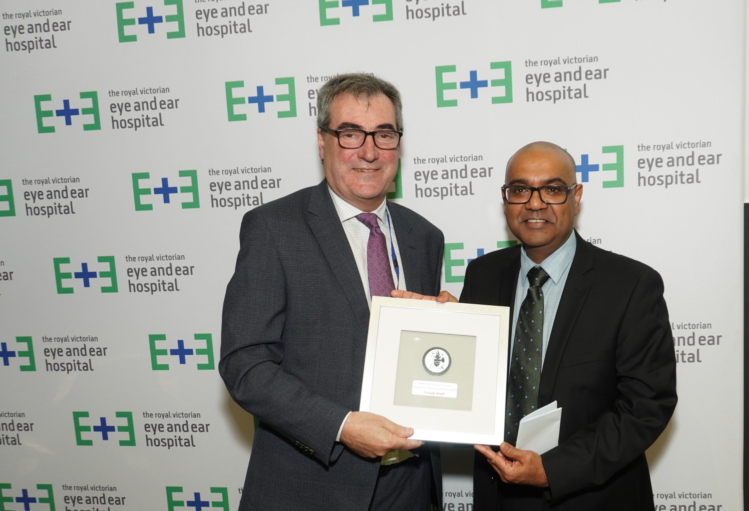 Executive Director Danny Mennuni presenting the Administrative Excellence Award to Sanjay Singh. They stand together smiling against an Eye and Ear logo background