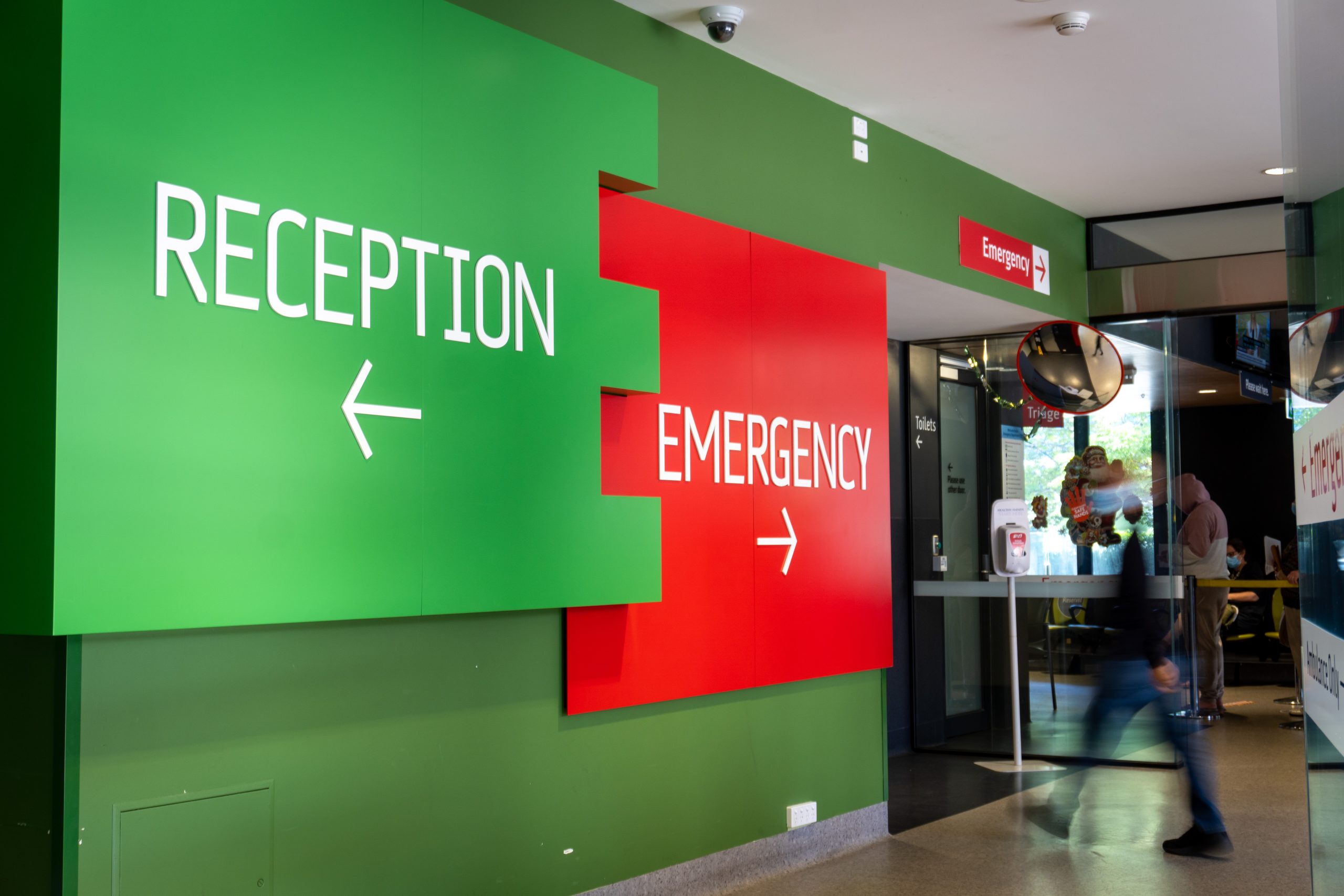 Image of the signage as you walk in from our Morrison Place entrance. A large green sign with white writing and an arrow pointing left reads 'Reception' and a red panel with white writing and an arrow pointing right says 'Emergency'.