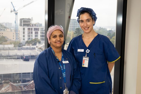 Nurse Smitha standing to the left of a graduate nurse, both wearing blue scrubs against a window