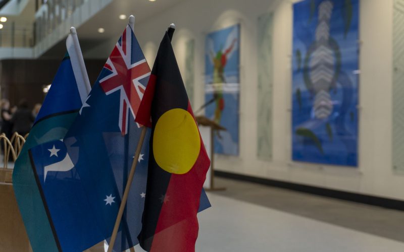 The Aboriginal, Torres Strait Islander and Australian flags hang in front of two large pieces of Aboriginal artwork.