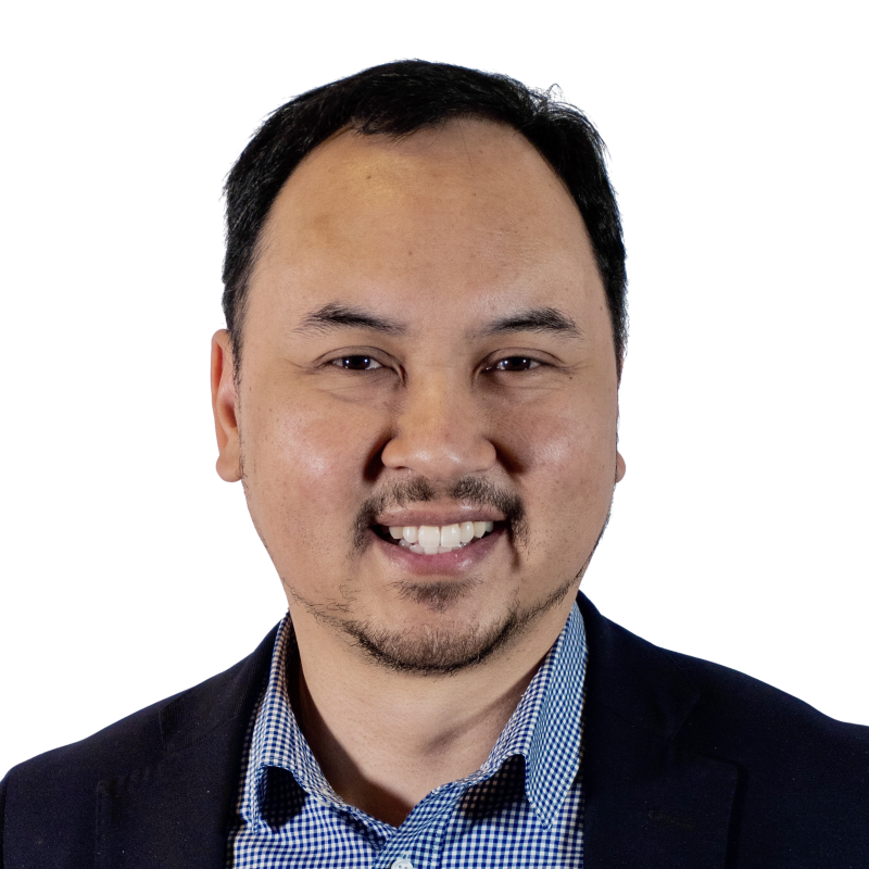 Dr Chris Tan, a man with short black hair wearing a black suit jacket and blue and white checkered shirt is smiling at the camera against a white background.