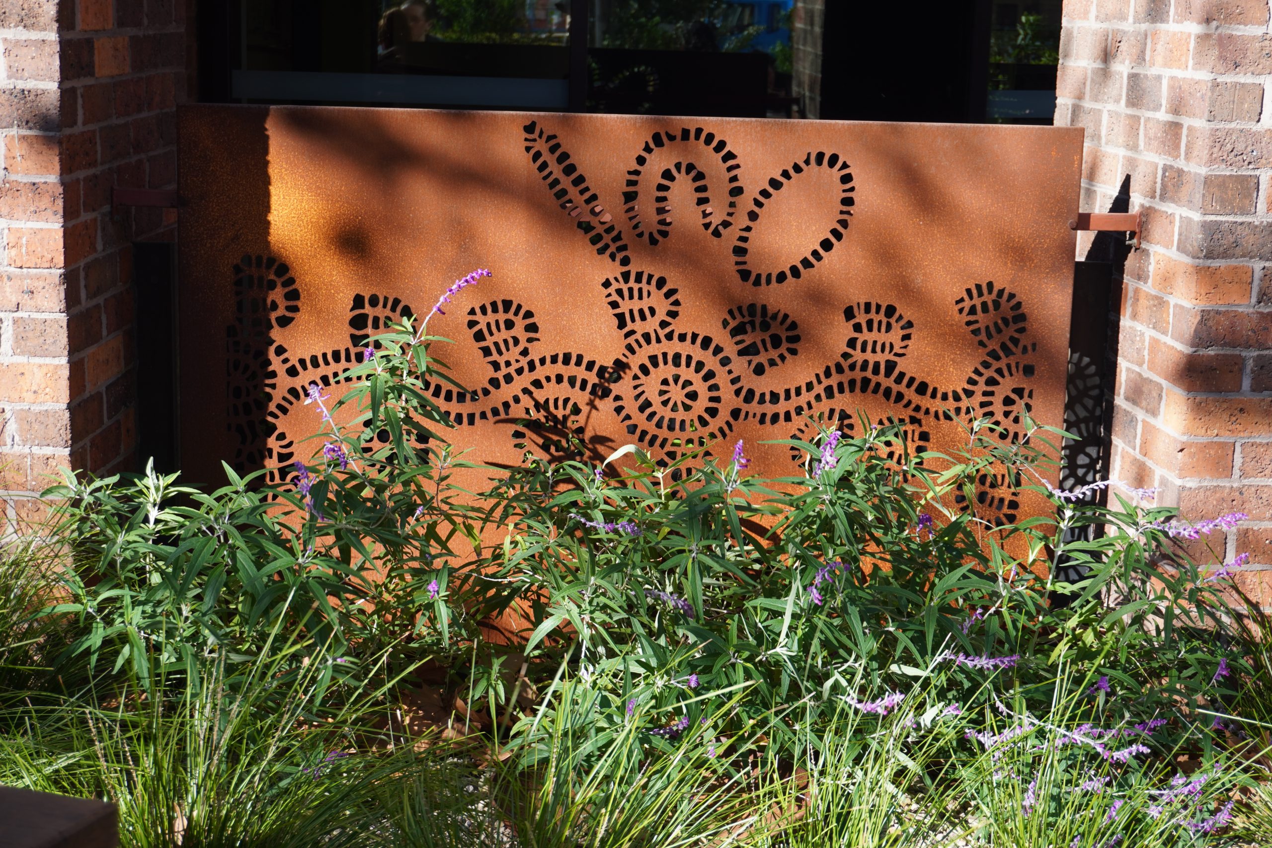 Photo of rusted panels commissioned for our Welcome Space. The rusted panels feature cut outs of Aboriginal art symbols and sit behind a native flower bed with purple flowers.