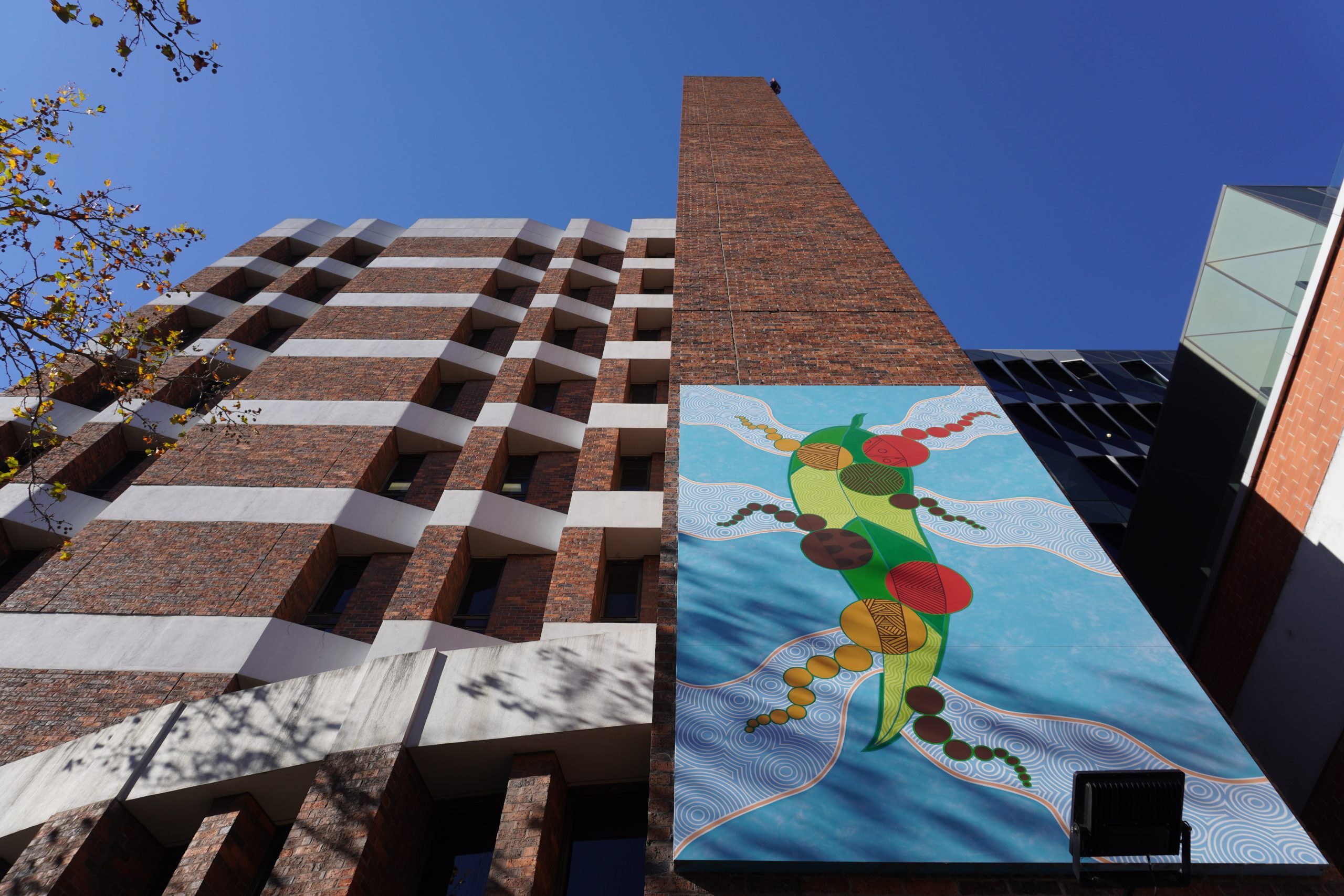 A photo looking up at Mandy Nicholson's Mannagum leaf mural on the side of the hospital above our Welcome Space against brown brick and a blue sky. The mural features a green mannagum leaf against a blue background with a number of Aboriginal art motifs in earthy colours.