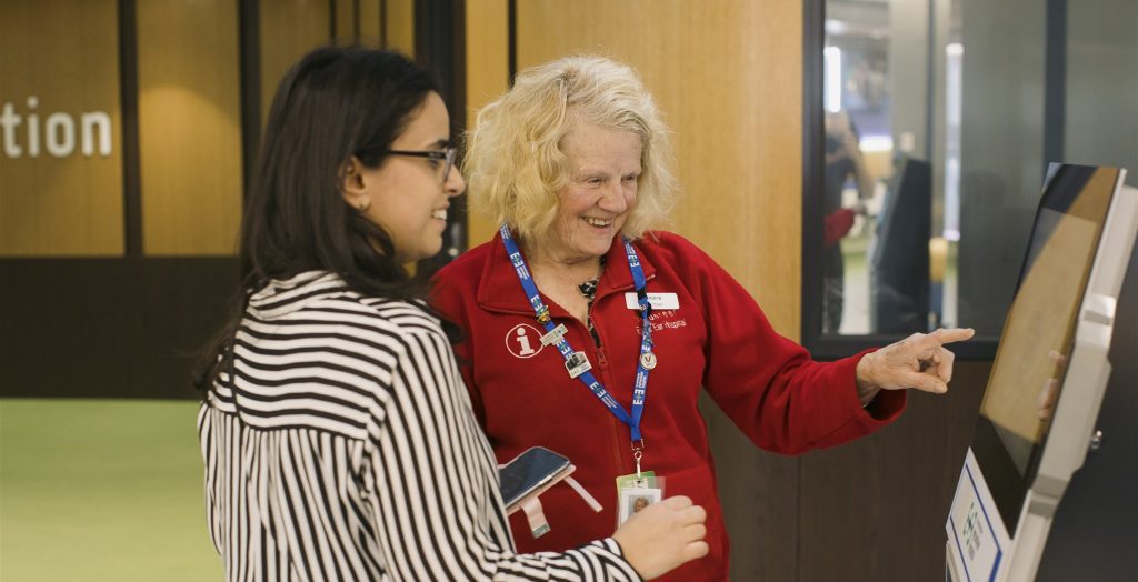 A volunteer in a red vest aiding a woman at the self check-in kiosk.