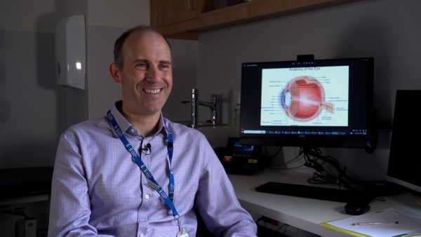 A man in a collared shirt sits in front of a computer with a diagram of an eye