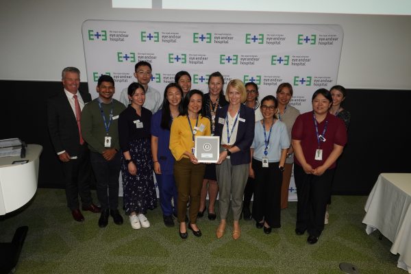 14 people stand together with and Eye and Ear banner in the background, holding an award