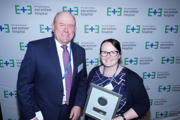 A man in a navy suit standing next to a woman wearing a black cardigan over a blue and white striped top holding a framed plaque