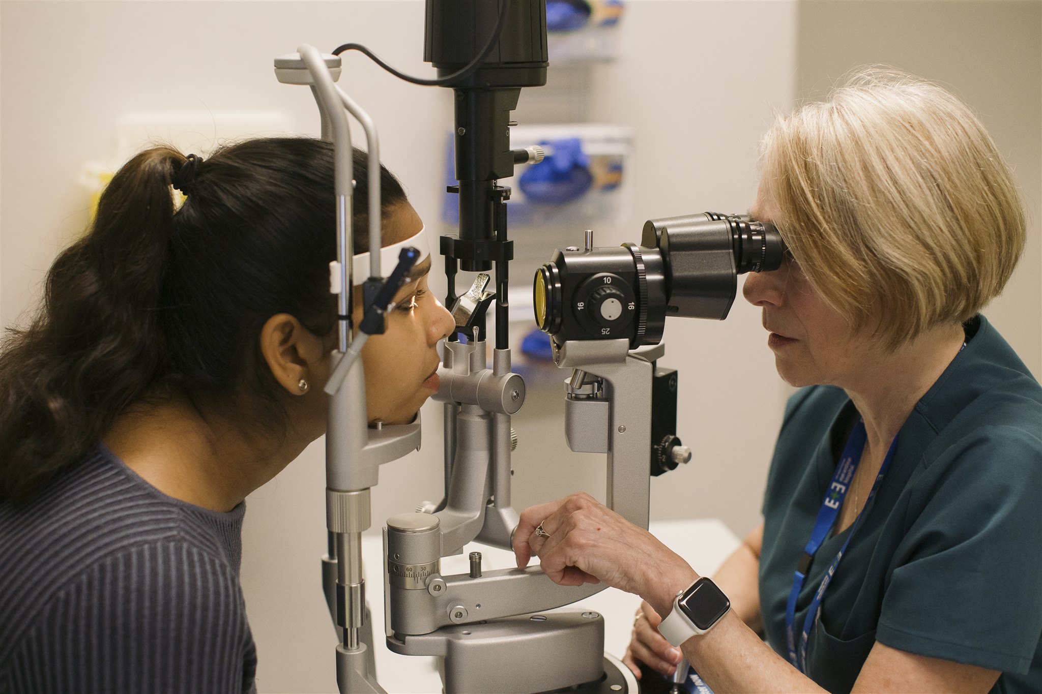 A patient sitting at a slit lamp have her eyes examined by a female in teal scrubs