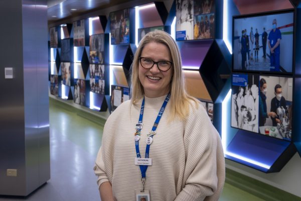 Catherine Mancuso standing in front of the colourful history wall in the foyer of the hospital. She wears a cream jumper, dark framed glasses and smiles at the camera