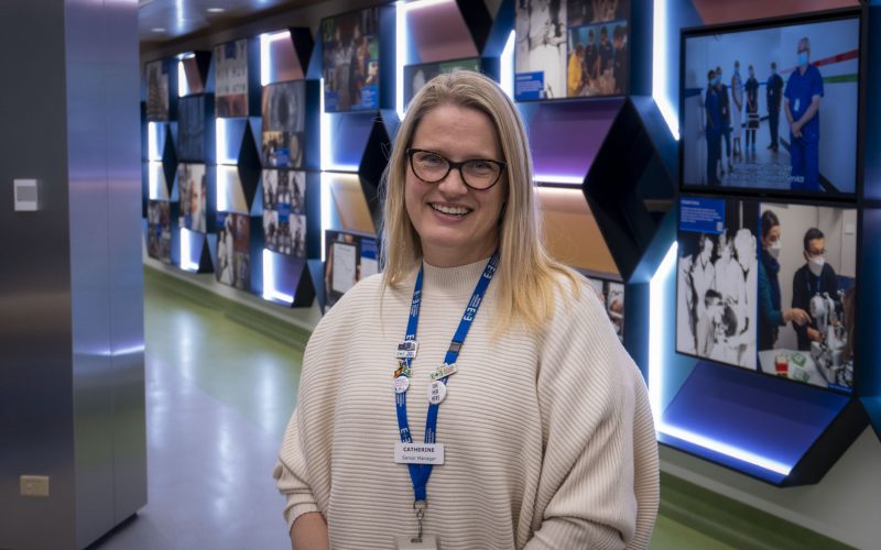 Catherine Mancuso standing in front of the colourful history wall in the foyer of the hospital. She wears a cream jumper, dark framed glasses and smiles at the camera