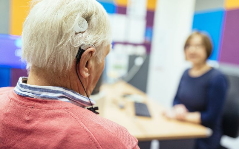 An older patient with a cochlear sits facing a health professional in a colourful room