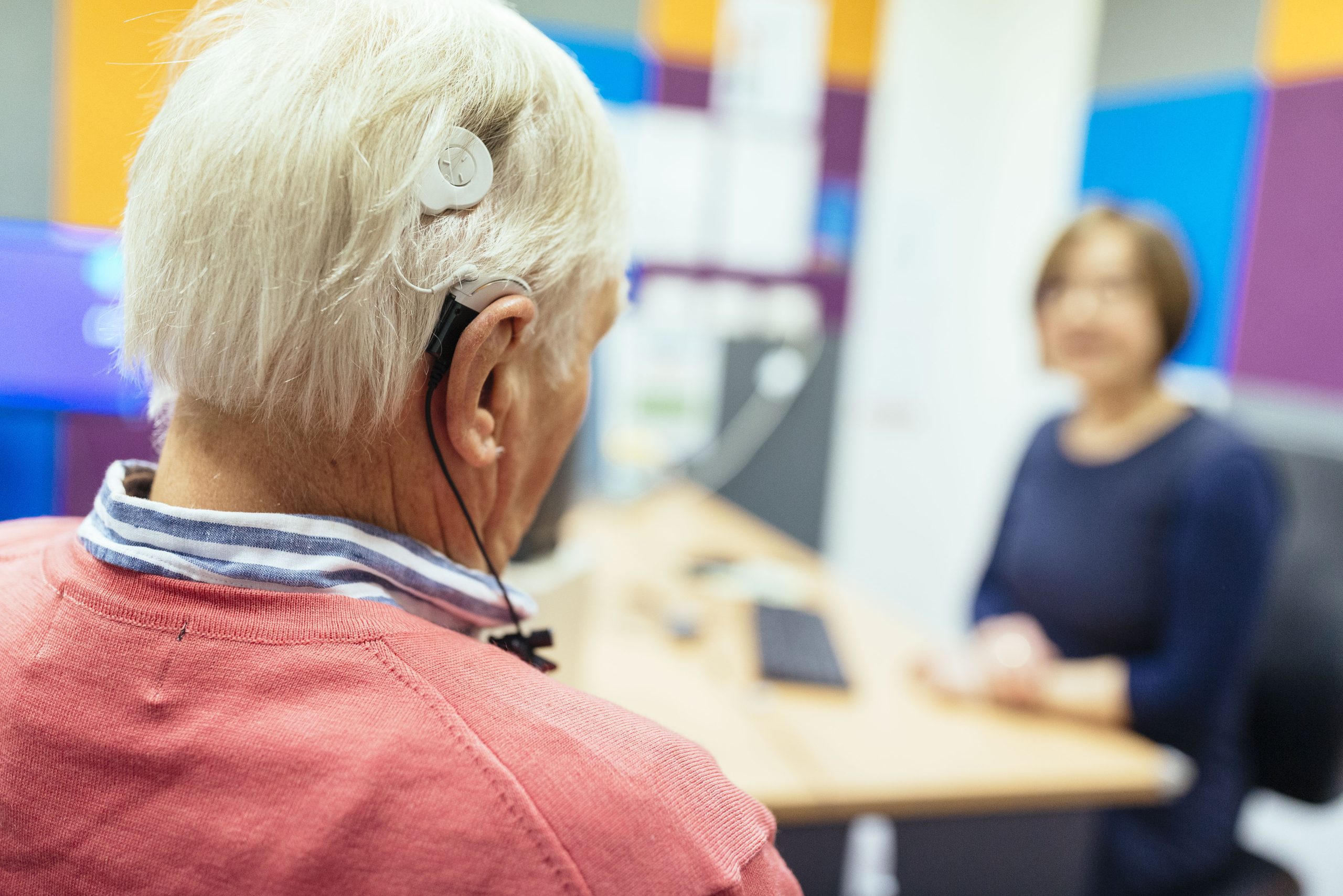 An older patient with a cochlear sits facing a health professional in a colourful room